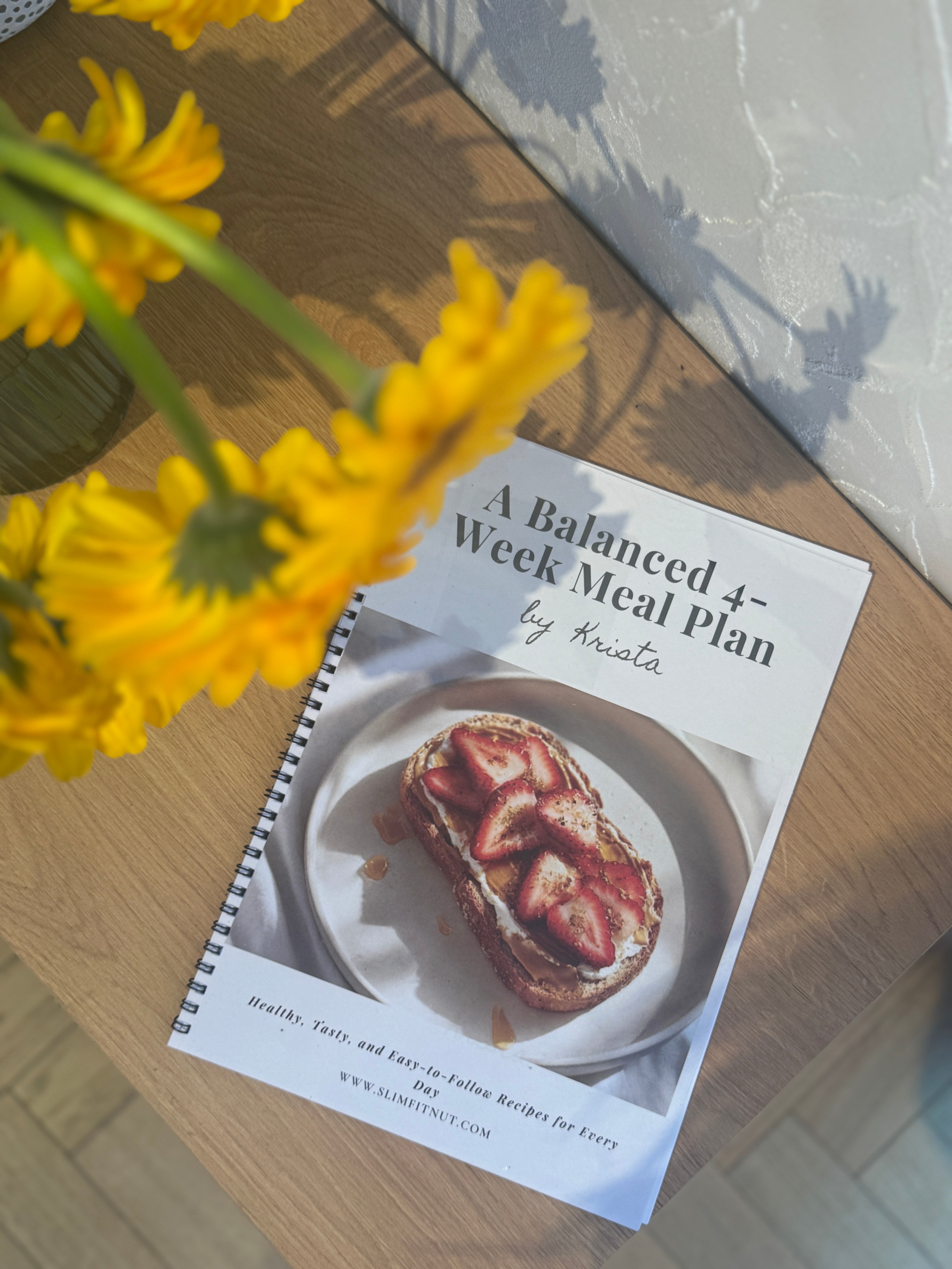 Printed ‘A Balanced 4-Week Meal Plan by Krista’ booklet on a wooden table, with a strawberry toast cover and yellow flowers casting soft shadows in natural light.